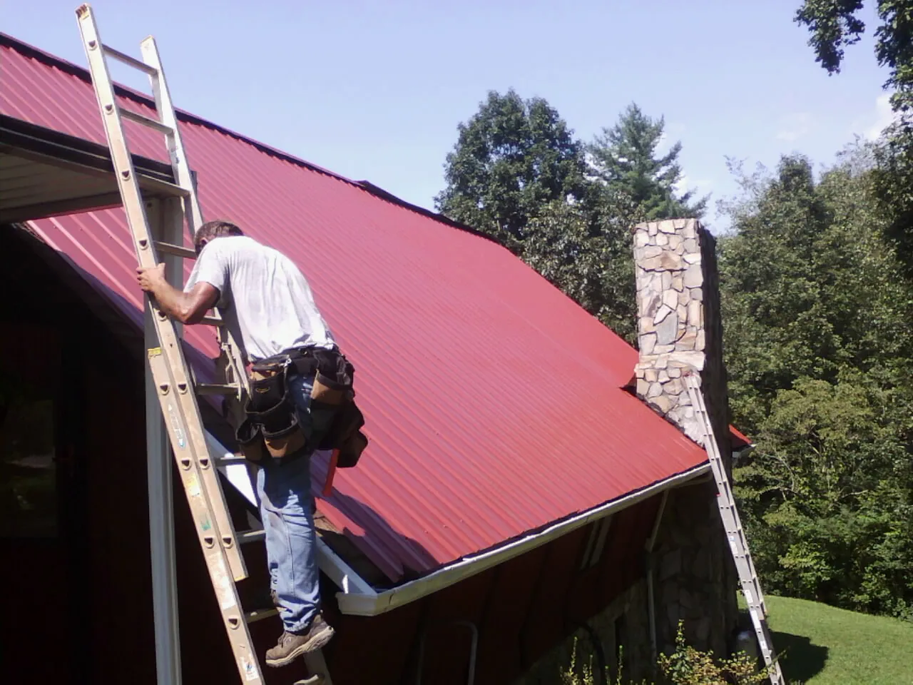 Roofer on a ladder beside a red metal roof and stone chimney — Soffit & Fascia Repair by Roofing Brothers in Newaygo